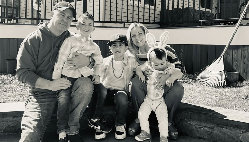 A family of five poses outdoors on a stone ledge. Two adults sit on either side, with three children between them. A rake is propped against the wall in the background.