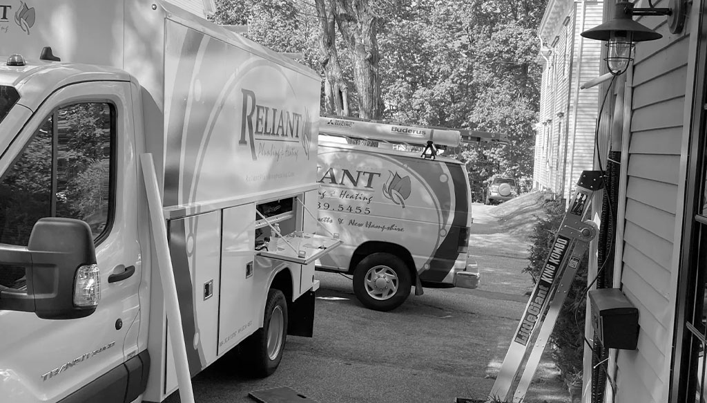 Two Reliant Heating and Air Conditioning service vans are parked outside a residential home. Ladders and equipment are visible nearby.