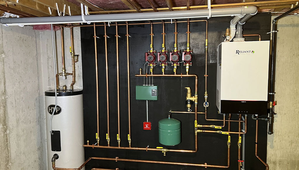 Image of a utility room shows a water heater tank, multiple copper pipes, valves, a green pressure tank, and a white heating unit mounted on the wall.