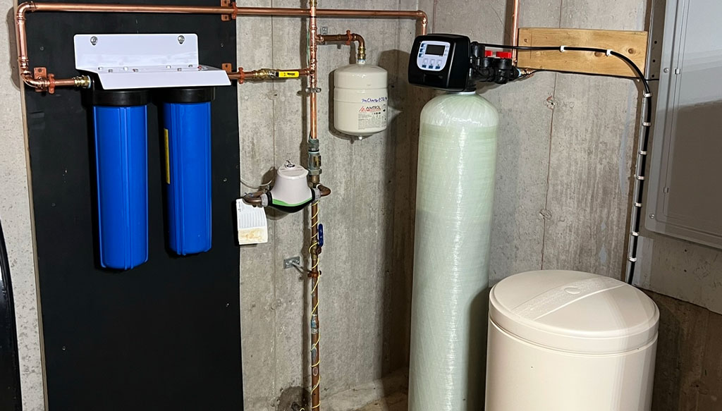 A water filtration system with blue filter cartridges and a control unit is installed on a wall. A large cylindrical tank and a white container are positioned next to it. Copper piping is visible.