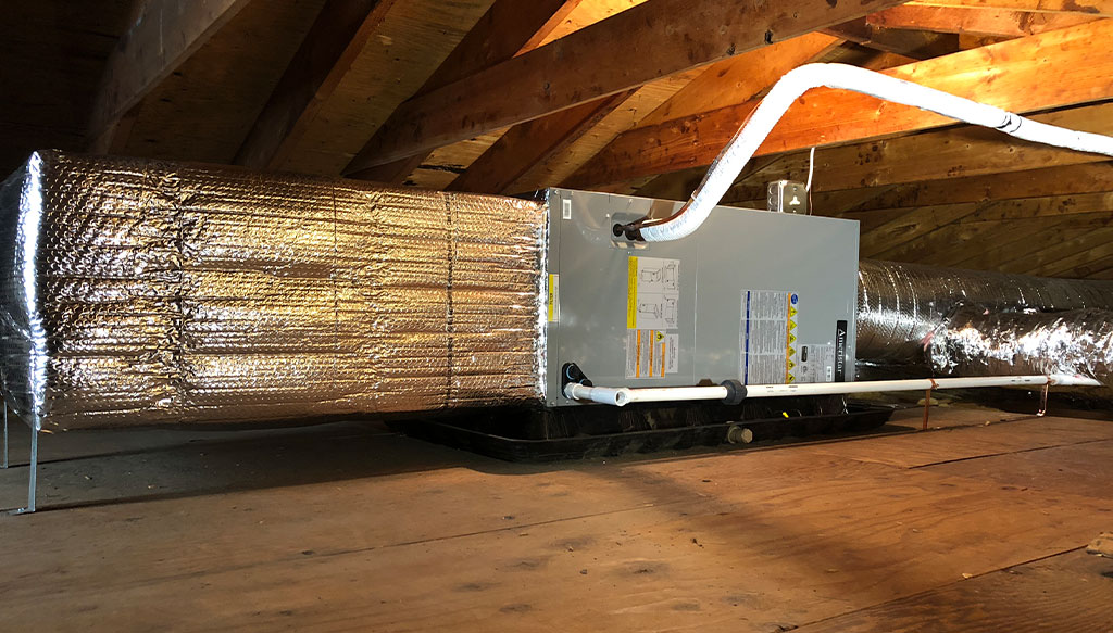 An HVAC unit installed in an attic with ductwork wrapped in reflective insulation and wooden beams overhead.