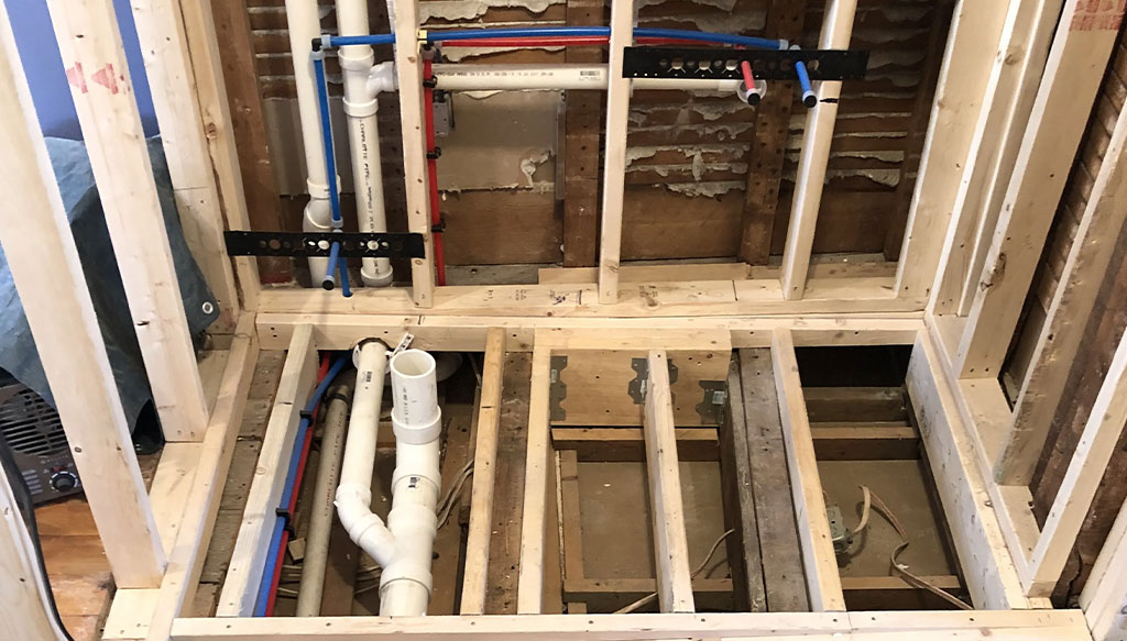 Exposed framing and plumbing in a house under construction, showing PVC and PEX pipes installed within wall and floor cavities.